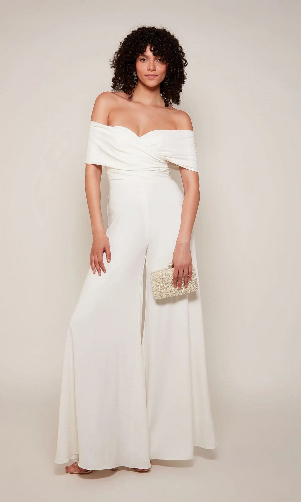 Woman wearing the Alyce 70051 long white jumpsuit with off-the-shoulder neckline and wide legs, holding a textured beige clutch against a plain light background.